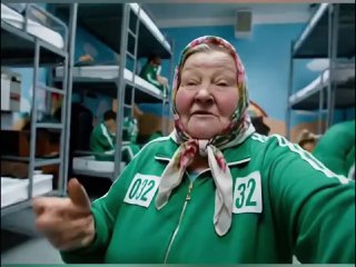 Пожилая женщина в платке и зелёном спортивном костюме с номером 032, на фоне спальных мест и людей в аналогичной форме, интерьер кадра.