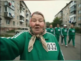 Та же пожилая женщина в зелёном костюме с номером 032 идёт по улице, рядом группа людей в одинаковой форме и жилые дома на фоне.
