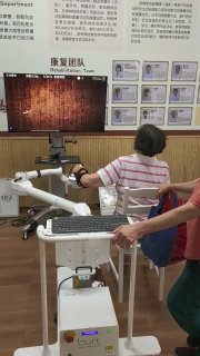 Пожилая пациентка в клинике сидит перед консолью и тренирует руку с помощью медицинского роботизированного устройства.