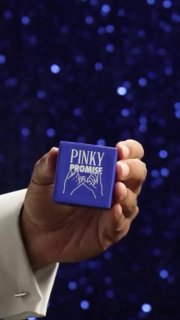 Рука держит маленькую фиолетовую коробочку с надписью «Pinky Promise» на фоне размытых синих огней; упаковка гаджета в кадре, демонстрируется продукт.