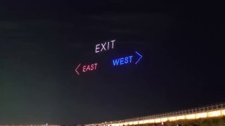 Ночное фото: дроны с подсветкой формируют в небе слово «EXIT» и направляющие стрелки «EAST» и «WEST» над местом мероприятия.