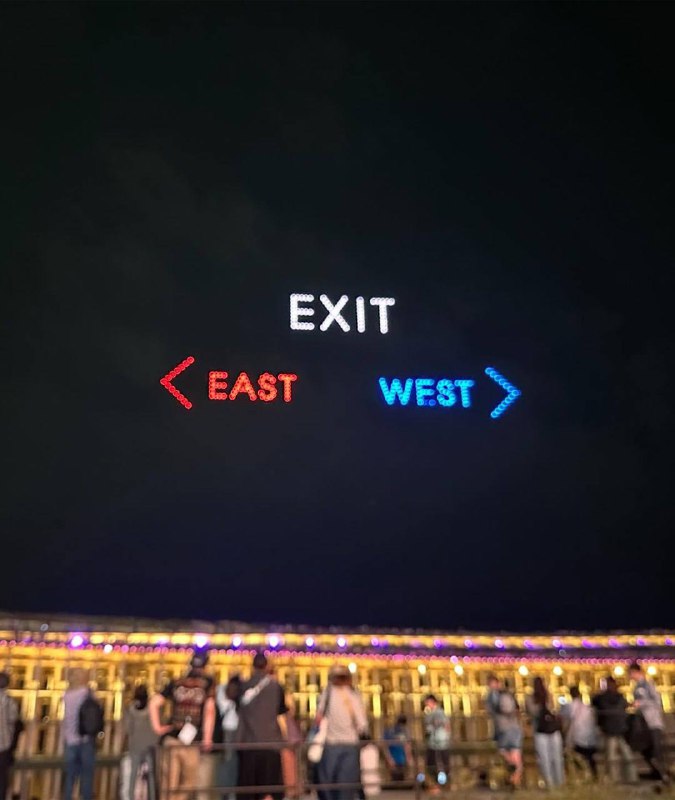 Крупный план ночного неба: яркие светящиеся стрелки и надпись EXIT, созданные дронами, на фоне огней и людей внизу.
