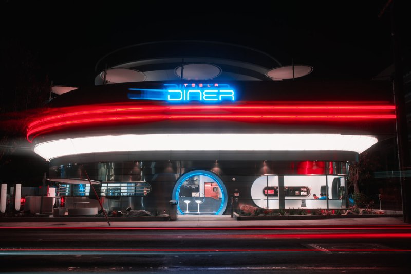 Ночной фасад Tesla Diner с яркой неоновой вывеской DINER, круглым входом и витриной; атмосфера ретро и современные элементы сервиса.