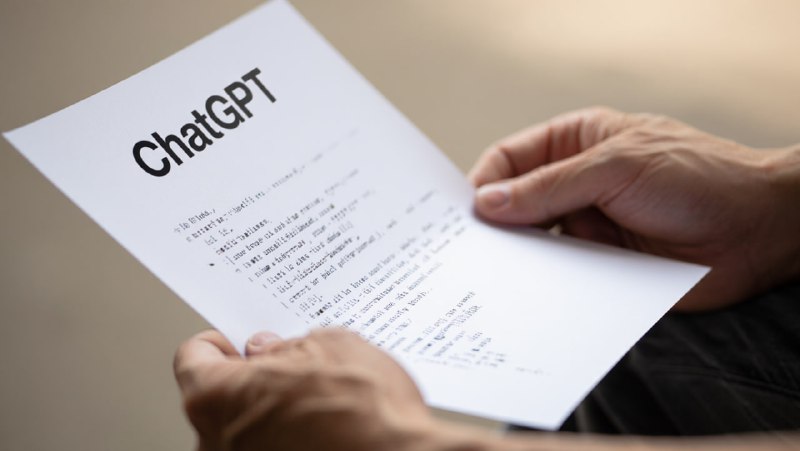 Человек держит в руках распечатанный лист с заголовком «ChatGPT» и списком подсказок; размытый фон, иллюстративная сцена с фокусом на бумаге и руках.