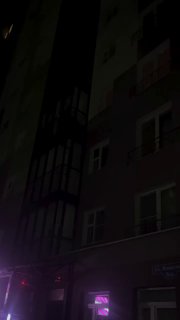 Ночное фото фасада жилого дома с фиолетовой подсветкой у входа; вид со двора, видно несколько окон, балконов и тёмные этажи.