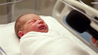Новорожденный, завернутый в одеяло в родильном боксе; рядом направлен микрофон, создавая контрастный образ «интервью» в роддоме.