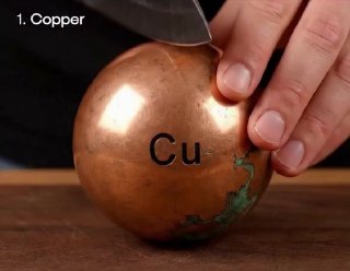 Рука с ножом рядом с медным шаром, на поверхности которого видна маркировка «Cu» и подпись «1. Copper», демонстрация металла.