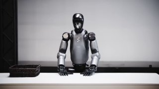 Гуманоидный робот Figure в студии, крупный план торса и рук-манипуляторов; на столе видна рабочая поверхность и корзина.