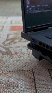 Крупный план боковой части ноутбука на ковре: виден USB‑порт и частично вставляемый штекер, ракурс снизу, бытовая обстановка.