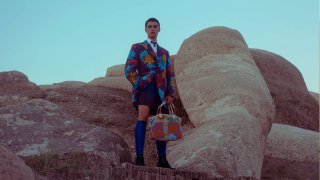 Человек в ярком костюме стоит на каменистом холме с сумкой; на заднем плане крупные скальные образования и небо.