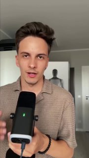 Молодой человек в комнате держит микрофон и говорит в камеру; вертикальное видео, на заднем плане дверь и силуэт объекта.