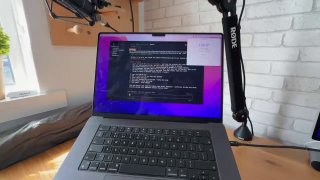 Фото рабочего места: MacBook с терминалом на экране, микрофон на стойке, деревянный стол и кирпичная стена на заднем плане.