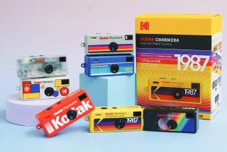Крупный план нескольких миниатюрных камер-брелоков Kodak Charmera с яркими коробками, аксессуарами и ретро-элементами на пастельном фоне.