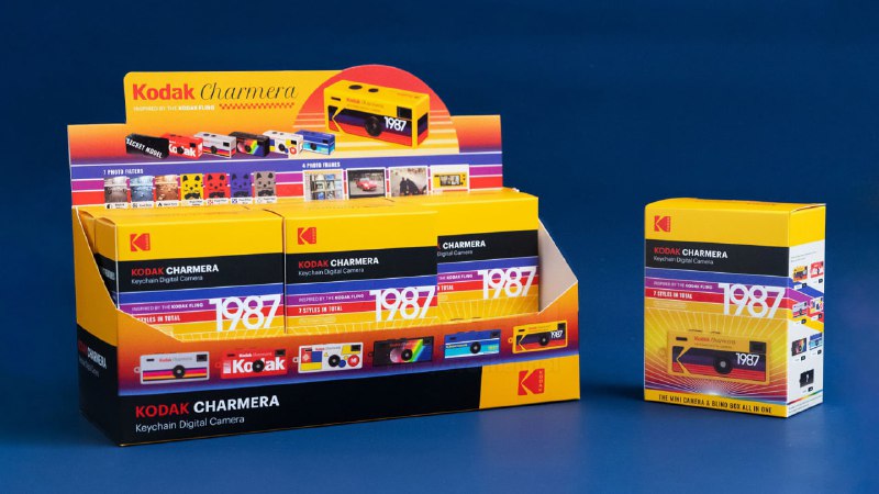 Выставочный дисплей с множеством коробок Kodak Charmera, показывающий ассортимент мини-камер-брелоков и их упаковку на синем фоне.
