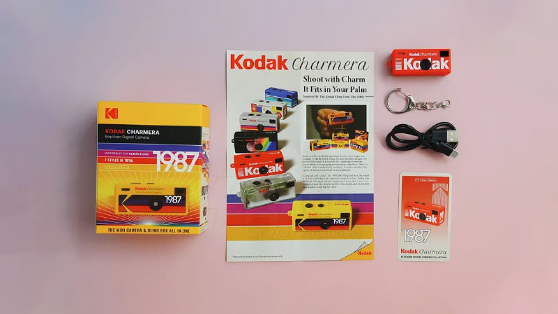 Композиция из коробки, листовки и нескольких брелок-камер Kodak Charmera с аксессуарами, представленных крупным планом на пастельной поверхности.