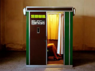 Фотокабина с зелёным корпусом и открытой жёлтой шторой; внутри видны ноги сидящего человека в туфлях, фон в тёплых тонах.