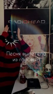 Комнатная фотография с плакатом Radiohead на стене; человек в полосатом свитере показывает на плакат, на столе предметы и декор.