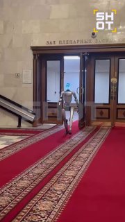 Гуманоидный робот идёт по длинному красному ковру в официальном зале здания к двойным дверям; видно интерьер и элементы зала.