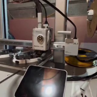 Крупный план рабочего стола с 3D-принтером: печатающая головка, катушка филамента и отражение на поверхности телефона.