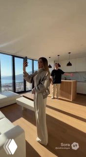 Женщина делает селфи в светлой квартире с панорамными окнами и видом на море; мужчина на фоне в минималистичном интерьере.