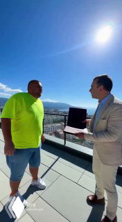 Два мужчины на открытой террасе с видом на море: один в неоновой футболке, другой в светлом костюме, обсуждают объект недвижимости на балконе при ярком солнце.