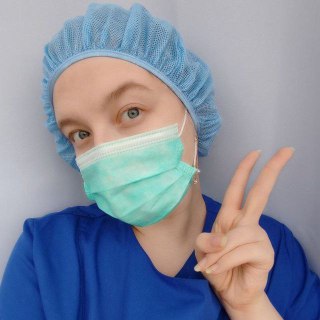 Портрет доулы Алины в медицинской форме и шапочке, в маске, показывает жест «V», нейтральный фон, дружелюбный и профессиональный образ.