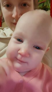 Селфи матери и младенца: крупный план лица малыша на переднем плане и частично видна мама на заднем плане в домашней обстановке, отражает повседневную заботу.