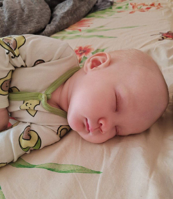 Спящий младенец в светлой пижаме, крупный план на лицо и плечи; спокойный дневной сон малыша на домашней кровати в уютной обстановке.