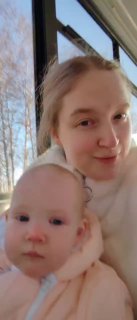 Мама с младенцем у окна в светлой домашней одежде, крупный план: лицо ребёнка и матери, за окном видны голые деревья и зимний пейзаж.