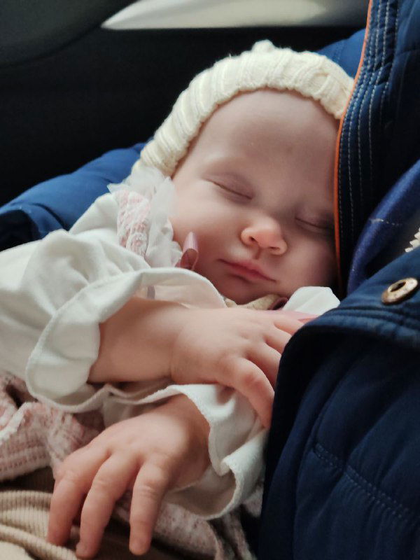 Спящий младенец в шапочке и тёплой одежде, прижатый к взрослому в прогулочной или автомобильной обстановке, спокойный и уютный, иллюстрация ношения и привязанности матери и ребёнка.