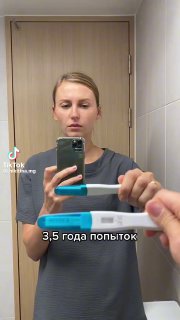 Женщина делает зеркальное селфи с телефоном и держит тест на беременность перед зеркалом; на кадре видна надпись «3.5 года попыток» и интерьер ванной комнаты.