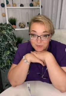 Доула Алина Морозова в фиолетовой рубашке и очках сидит за столом, улыбаясь в камеру на фоне полок и растений в уютной комнате.