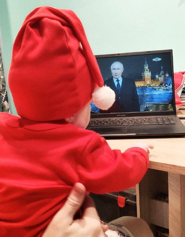 Ребёнок в красном костюме сидит перед ноутбуком и внимательно смотрит трансляцию речи президента на экране в домашней обстановке, кадр в интерьере.