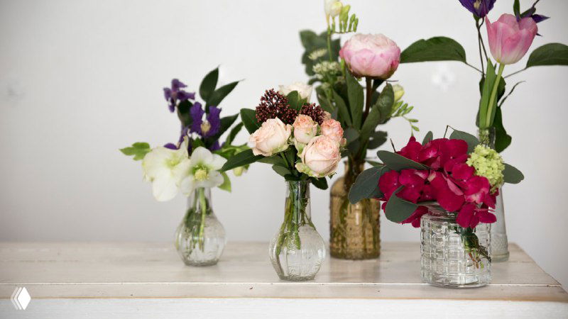 Несколько небольших букетов в стеклянных вазах на белой полке — примеры оформления и хранения срезанных цветов в домашних условиях.