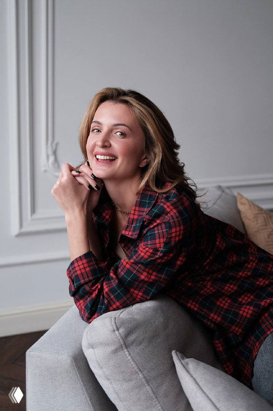 Женщина в клетчатой рубашке улыбается, сидя на диване в уютной светлой комнате — портрет бизнес-медиатора в неформальной обстановке.