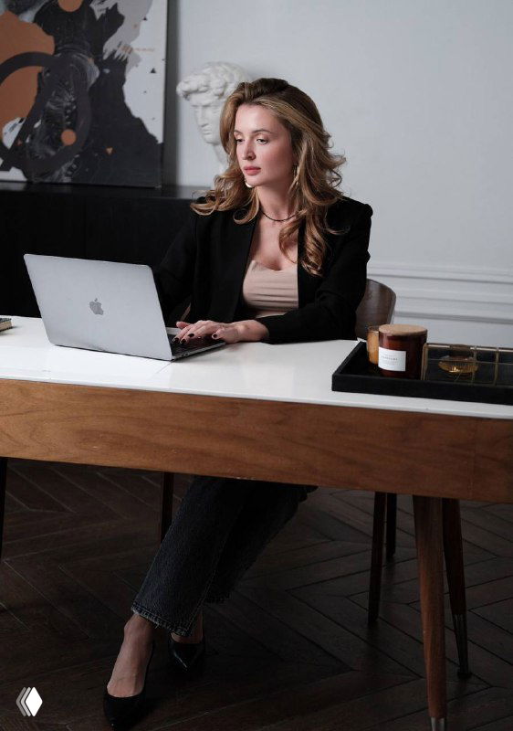 Женщина в деловой одежде сидит за деревянным столом с открытым ноутбуком, работает в уютном офисе рядом со свечой и подносом.