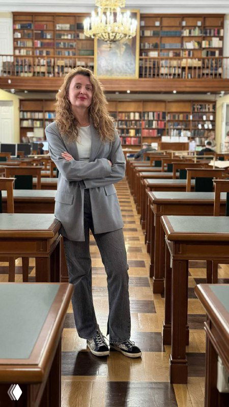 Женщина в деловом костюме стоит в читальном зале библиотеки между деревянными столами; образ создает ассоциацию с медиатором или консультантом.