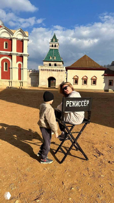 Человек в раскладном кресле с надписью «РЕЖИССЁР» беседует с ребёнком на фоне старинного здания и ясного голубого неба.