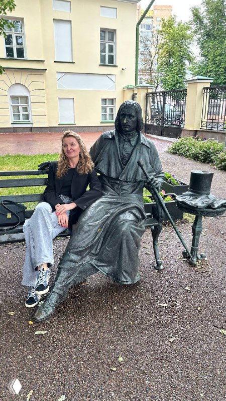 Женщина сидит на скамейке рядом с бронзовой статуей человека в парке у здания, поза показывает дружелюбную сцену на улице летом.