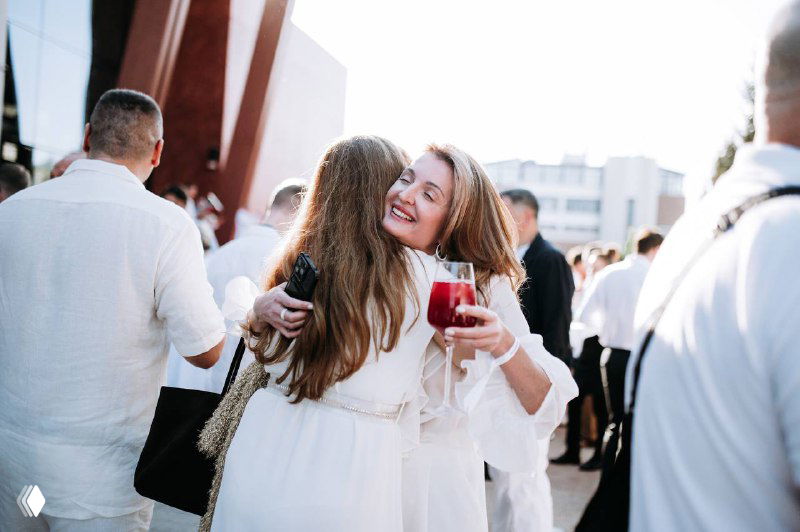 Деловой приём на открытом воздухе: люди в светлой одежде обнимаются и общаются, женщина держит бокал вина, атмосфера нетворкинга.