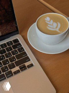 Крупный план рабочего места: кружка капучино с латте‑артом на белом блюдце рядом с клавиатурой ноутбука на деревянном столе, деловая атмосфера.