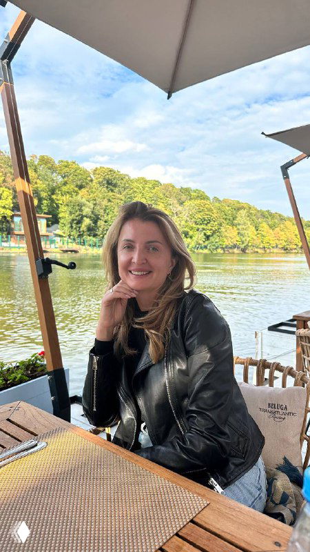 Женщина улыбается за столиком на летней веранде у реки, в кожаной куртке; на заднем плане зелёные берега и зонты.