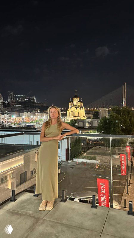 Женщина в длинном платье стоит на террасе ночью; на заднем плане подсвеченные здания, мост и городские улицы.