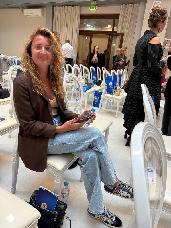 Женщина в пиджаке и джинсах сидит на стуле в светлом зале с телефоном в руках; кадр показывает атмосферу деловой встречи.