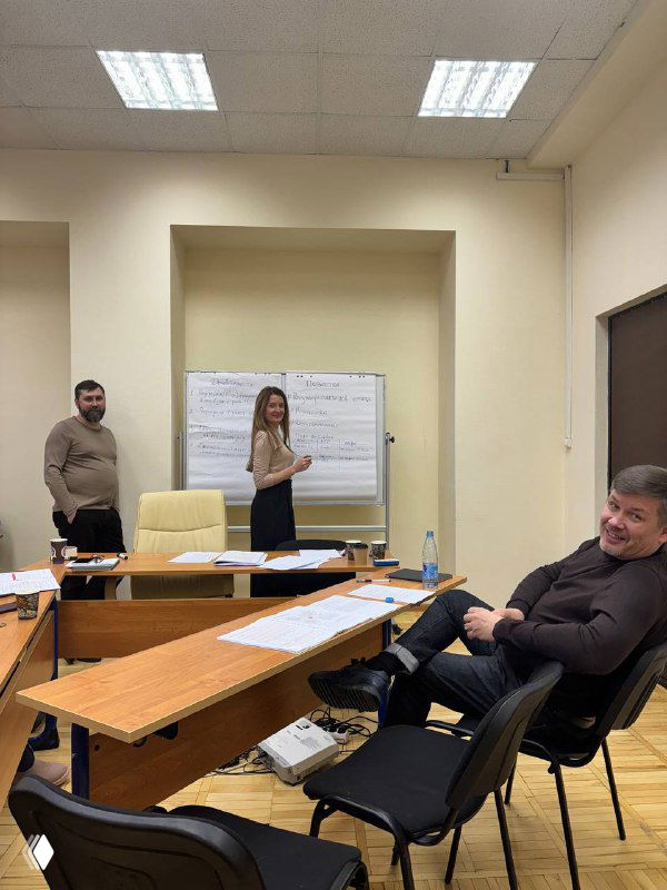 Переговоры в офисе: три участника за столом, флипчарт с заметками на стене, документы и ноутбук в кадре.