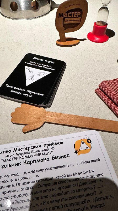 Крупный план игровых элементов: деревянный указатель, колода карт и фрагмент инструкции, используемые в тренинге по распознаванию ролей жертвы, агрессора и спасателя.