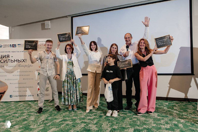 Сцена конференции по медиации: группа участников поднимает руки и получает награды на фоне проекционного экрана и брендированного баннера.