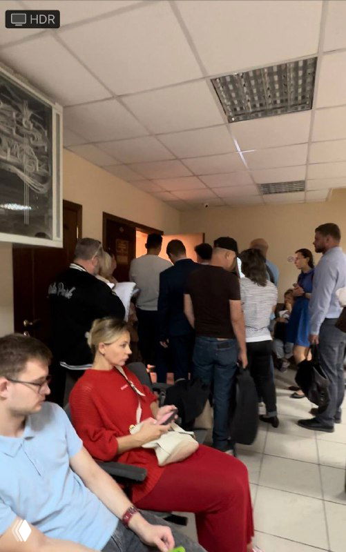 Группа людей в коридоре суда, ожидающих начала заседания: стоят и сидят, ощущается напряжённая атмосфера ожидания и недовольства.