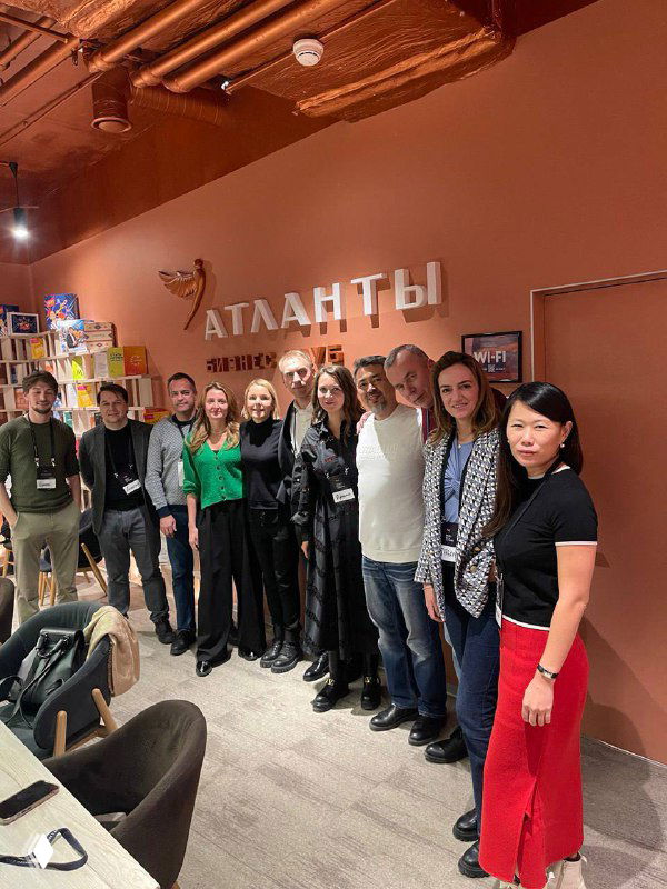 Групповое фото участников и организаторов в интерьере Бизнес‑клуба «Атланты»: люди стоят у стены с логотипом клуба, улыбающиеся участники с бейджами.