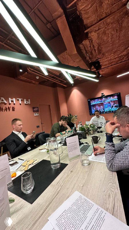 Участники сидят за столом во время управленческого поединка: обсуждения, заметки, бокалы воды и бланки с кейсами на столе, живая рабочая сессия.